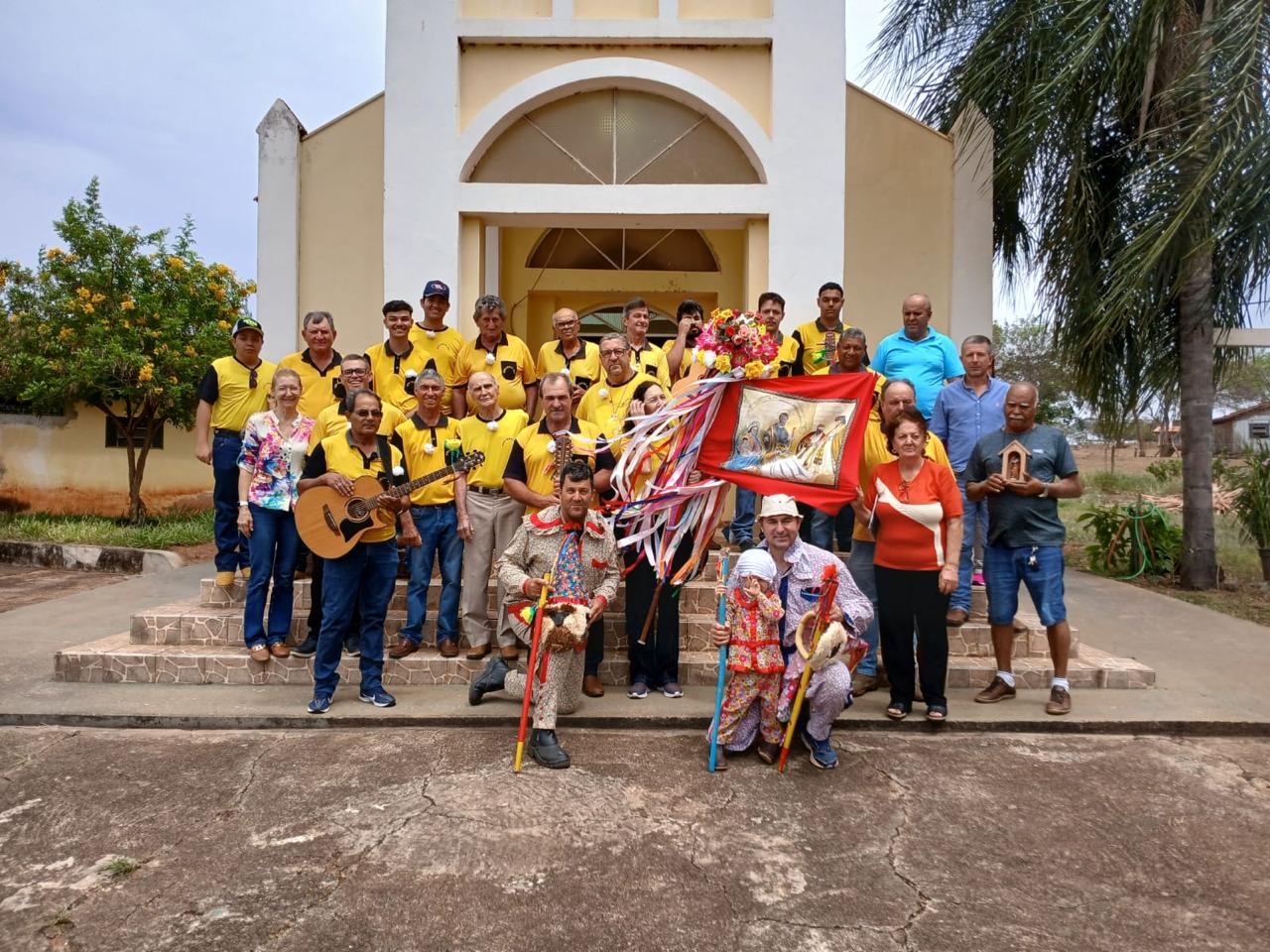 Companhia de Santos Reis do Bairro do Campinho reunida em frente à Igreja de São Benedito, com integrantes em camisetas amarelas, estandarte e palhaços da folia